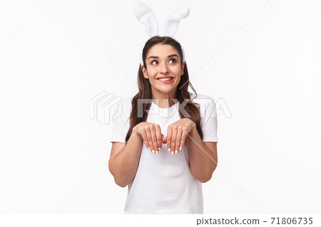 Portrait of funny, lovely european girl with rabbit ears, imitating bunny, holding hands like paws and looking up daydreaming, imaging something cute, celebrating Easter day, white background Portrait of funny, lovely european girl with rabbit ears, imitating bunny, holding hands like paws and looking up daydreaming, imaging something cute, celebrating Easter day, white background 71806735