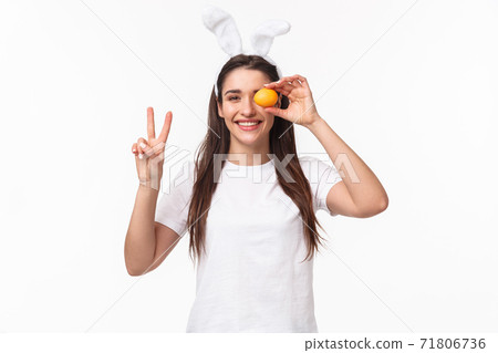 Portrait of peaceful and cheerful young cute female in white t-shirt, showint peace sign and hold colored egg over eye, smiling happy, enjoying traditional holiday celebration, Easter day Portrait of peaceful and cheerful young cute female in white t-shirt, showint peace sign and hold colored egg over eye, smiling happy, enjoying traditional holiday celebration, Easter day 71806736