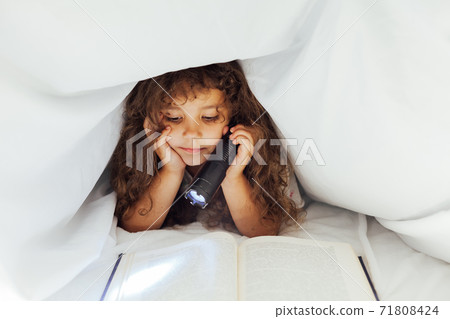 Little girl reads a book at night with a flashlight under a blanket Little girl reads a book at night with a flashlight under a blanket 71808424