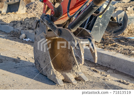 Close up of the metal bucket of a red excavator against a road lit by sunlight 71813519
