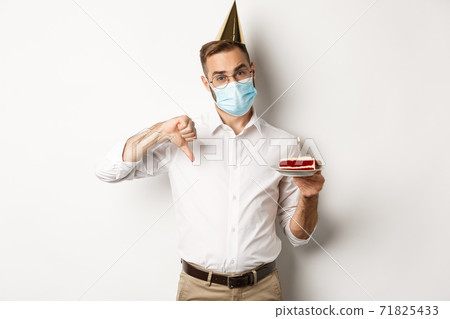 Coronavirus, quarantine and holidays. Man showing thumb down as disappointed with birthday party, wearing face mask and holding bday cake, white background 71825433