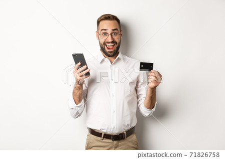 Business and online payment. Excited man paying with mobile phone and credit card, smiling amazed, standing over white background Business and online payment. Excited man paying with mobile phone and credit card, smiling amazed, standing over white background 71826758
