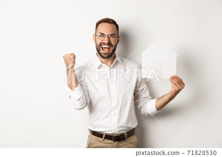 Real estate. Satisfied man rejoicing of founding perfect home apartment, holding paper house model, standing over white background Real estate. Satisfied man rejoicing of founding perfect home apartment, holding paper house model, standing over white background 71828530