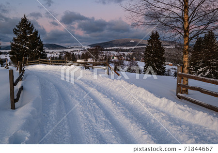 Snow drifts on road through twilight small and quiet winter alpine village, Voronenko, Carpathian, Ukraine. Night countryside hills, groves and farmlands in remote alpine village. 71844667