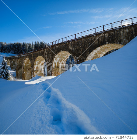 Stone viaduct (arch bridge) on railway through mountain snowy fir forest. Snow drifts  on wayside and hoarfrost on trees. 71844732