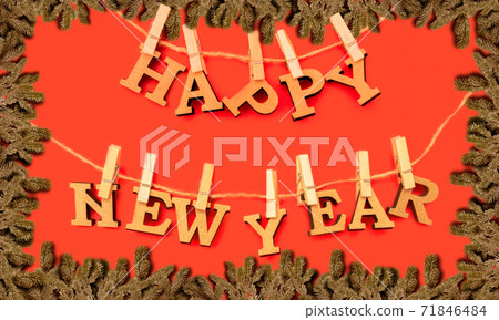 Happy New Year. Wooden letters in a New Year's cap on a red background with fir branches. Holiday decoration or postcard concept with top view and copy space Happy New Year. Wooden letters in a New Year's cap on a red background with fir branches. Holiday decoration or postcard concept with top view and copy space 71846484