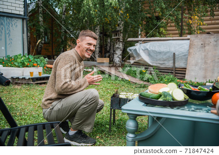 A young man in the backyard of the garden roasts sliced zucchini on the grill in the grill, next to a table with other vegetables A young man in the backyard of the garden roasts sliced zucchini on the grill in the grill, next to a table with other vegetables 71847424