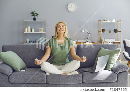 Smiling relaxed business woman meditating while working at home sitting near laptop on sofa. Smiling relaxed business woman meditating while working at home sitting near laptop on sofa. 71847865