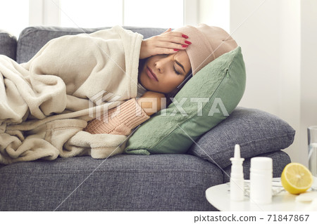 Close up of a sick woman with flu, fever and headache lying wrapped in a plaid on the couch. Close up of a sick woman with flu, fever and headache lying wrapped in a plaid on the couch. 71847867