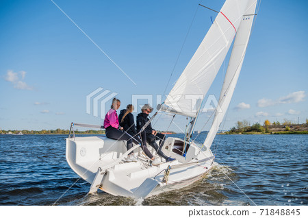 A beautiful white racing single-masted yacht is sailing against a beautiful river landscape with a blue sky. A man and two girls are on board. A beautiful white racing single-masted yacht is sailing against a beautiful river landscape with a blue sky. A man and two girls are on board. 71848845
