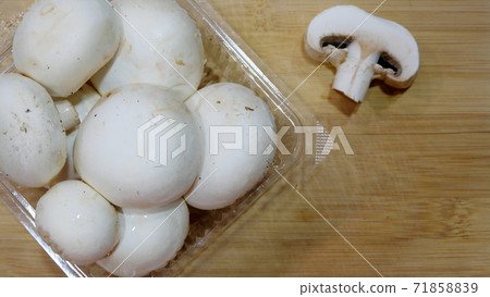 Close up of a transparent plastic package of white button mushrooms, with the lid opened, placed on top of a wooden cutting board. With a cut slice at the side. 71858839