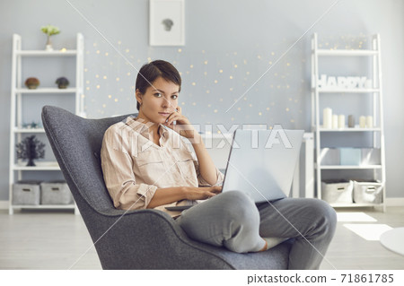 Serious woman looking at camera while working on laptop sitting in cozy armchair 71861785