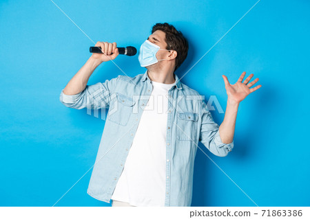 Handsome guy in medical mask singing in microphone, standing over blue background 71863386