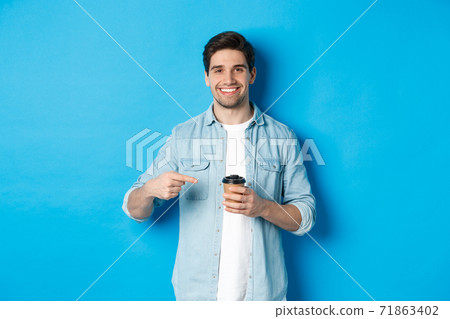 Smiling happy man pointing at paper cup with coffee, recommending cafe, standing over blue background Smiling happy man pointing at paper cup with coffee, recommending cafe, standing over blue background 71863402