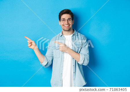 Young handsome man in glasses showing advertisement, smiling and pointing fingers left, making announcement, standing against blue background 71863405