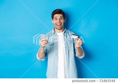 Amazed handsome man shopping online, holding mobile phone and credit card, smiling while paying for internet purchase, standing over blue background Amazed handsome man shopping online, holding mobile phone and credit card, smiling while paying for internet purchase, standing over blue background 71863548