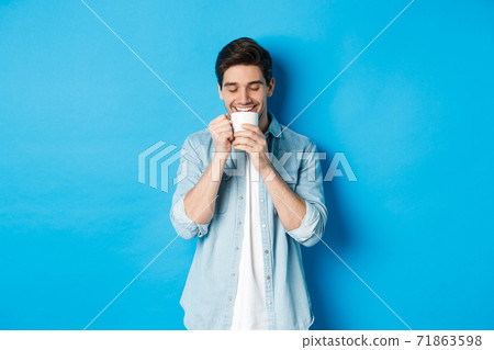 Young man drinking coffee with happy face, standing over blue background Young man drinking coffee with happy face, standing over blue background 71863598
