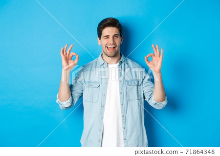 Relaxed and confident man showing ok signs and winking, everything okay gesture, standing against blue background Relaxed and confident man showing ok signs and winking, everything okay gesture, standing against blue background 71864348