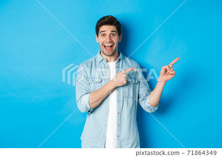 Image of excited handsome man in casual outfit, showing advertisement, pointing fingers right at copy space and smiling, standing against blue background Image of excited handsome man in casual outfit, showing advertisement, pointing fingers right at copy space and smiling, standing against blue background 71864349