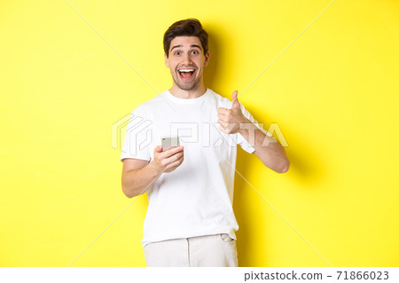 Happy smiling man holding smartphone, showing thumb up in approval, recommend something online, standing over yellow background 71866023