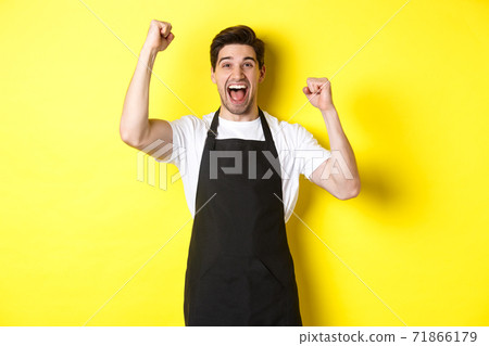 Happy barista celebrating victory, raising hands up and shouting for joy, wearing black apron, shop uniform, standing against yellow background Happy barista celebrating victory, raising hands up and shouting for joy, wearing black apron, shop uniform, standing against yellow background 71866179