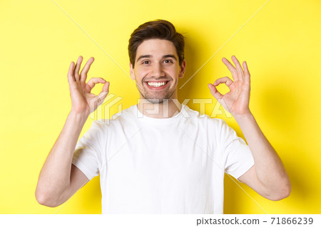 Close-up of handsome young man showing okay sign, approve and agree, smiling satisfied, standing over yellow background 71866239