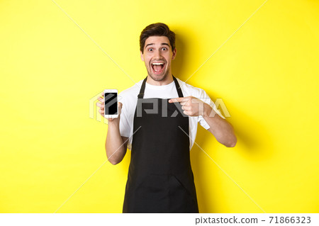 Handsome barista in black apron pointing finger at mobile screen, showing app and smiling, standing over yellow background Handsome barista in black apron pointing finger at mobile screen, showing app and smiling, standing over yellow background 71866323