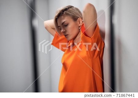 A beautiful blonde criminal in an orange robe stands in her cell behind bars, leaning against the wall 71867441