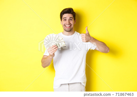 Satisfied buyer man showing thumb-up and holding money, shopping with cash, standing over yellow background Satisfied buyer man showing thumb-up and holding money, shopping with cash, standing over yellow background 71867463