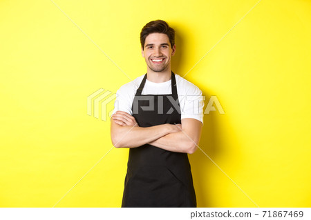 Smiling male waiter in black apron standing confident, cross arms on chest against yellow background 71867469