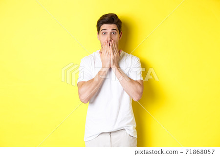 Handsome man looking shocked and speechless, holding hands on mouth, standing over yellow background Handsome man looking shocked and speechless, holding hands on mouth, standing over yellow background 71868057