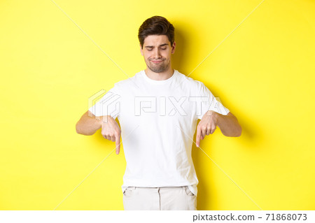Skeptical young man in white t-shirt, pointing and looking down upset, disapprove and dislike product, standing over yellow background 71868073