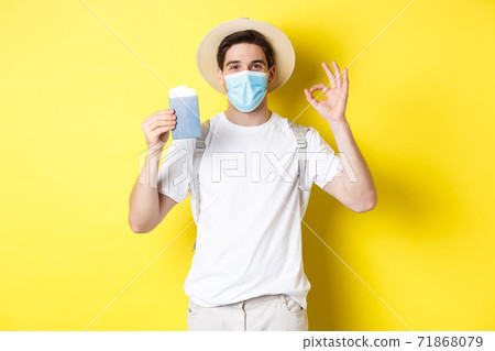 Concept of covid-19, tourism and pandemic. Happy male tourist in medical mask showing passport, going on vacation during coronavirus, make ok sign, yellow background 71868079