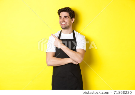 Handsome barista pointing and looking left at promo, wearing black apron, standing against yellow background 71868220