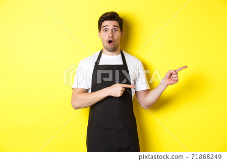 Amazed waiter in black apron showing promo offer, pointing fingers right and looking surprised, standing in uniform against yellow background 71868249