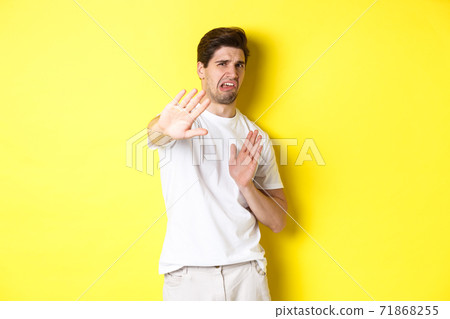 Disgusted man refusing, grimacing from dislike and aversion, begging to stop, standing in white t-shirt against yellow background Disgusted man refusing, grimacing from dislike and aversion, begging to stop, standing in white t-shirt against yellow background 71868255