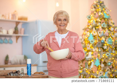 Grey-haired woman in pink shirt stirring something in a bowl and looking cheerful 71869111