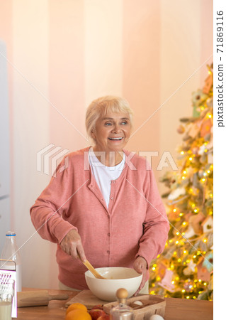 Smiling grey-haired woman cooking something in the kitchen and looking contented Smiling grey-haired woman cooking something in the kitchen and looking contented 71869116
