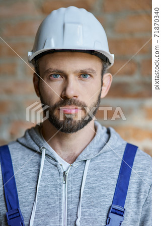 Close up portrait of confident attractive young male builder in hard hat looking at camera, posing on a brick wall background, working at construction site Close up portrait of confident attractive young male builder in hard hat looking at camera, posing on a brick wall background, working at construction site 71870340