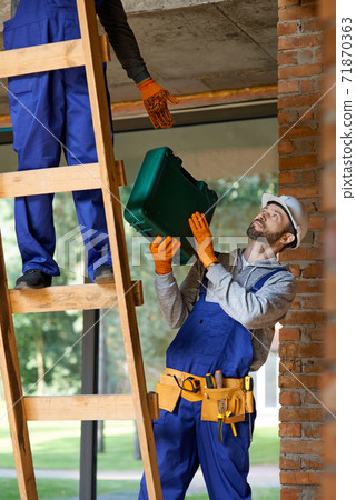 Young workman in overalls and hard hat giving toolbox to his colleague on ladder while working at cottage construction site Young workman in overalls and hard hat giving toolbox to his colleague on ladder while working at cottage construction site 71870363