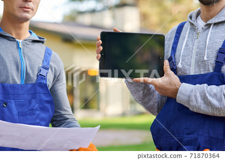 Cropped shot of young male builder showing screen of digital tablet pc with coworker holding blueprint in the background, working at cottage construction site Cropped shot of young male builder showing screen of digital tablet pc with coworker holding blueprint in the background, working at cottage construction site 71870364
