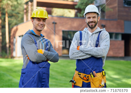 Experts in remodelling. Portrait of two handsome young male engineers in hard hats smiling at camera, posing outdoors with arms crossed while working on cottage construction Experts in remodelling. Portrait of two handsome young male engineers in hard hats smiling at camera, posing outdoors with arms crossed while working on cottage construction 71870370