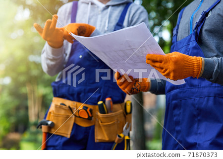 New project. Close up shot of two builders standing outdoors with an open blueprint discussing the cottage construction New project. Close up shot of two builders standing outdoors with an open blueprint discussing the cottage construction 71870373