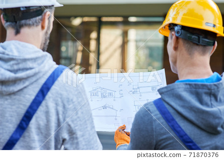 Plan better. Close up of two builders standing outdoors with an open blueprint discussing the interior of a half completed cottage house 71870376