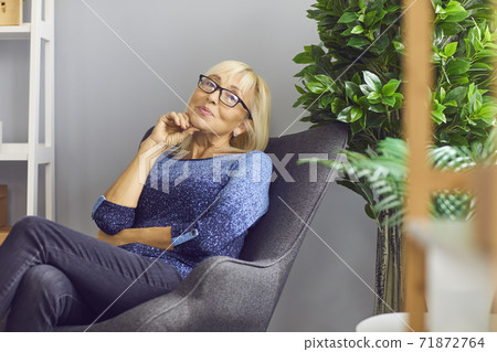 Independent mature woman sitting in armchair at home, thinking and looking at camera 71872764