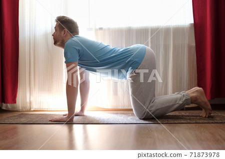 man practices yoga asana marjariasana or cow pose at home. 71873978