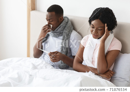 Sick african couple sitting in bed, measuring fever, sneezing noses 71876534