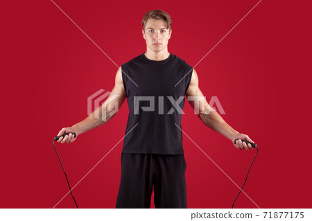 Aerobic workout concept. Serious athletic millennial guy jumping on skipping rope over red studio background Aerobic workout concept. Serious athletic millennial guy jumping on skipping rope over red studio background 71877175