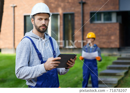 Portrait of young male builder looking at camera while holding digital tablet pc with coworker working in the background at cottage construction site Portrait of young male builder looking at camera while holding digital tablet pc with coworker working in the background at cottage construction site 71878207