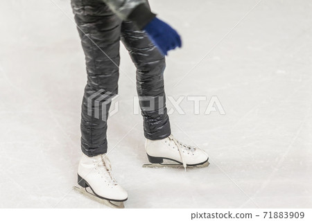 Unrecognizable girl in a black tracksuit skating on ice rink in city park, legs with skates close up. Snow and winter fun. Healthy outdoor activity. 71883909